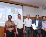 Fünf Männer neben einander stehend blicken in die kamera, im Hintergrund die Projektion weiterer Teilnehmer einer Online-Konferenz.