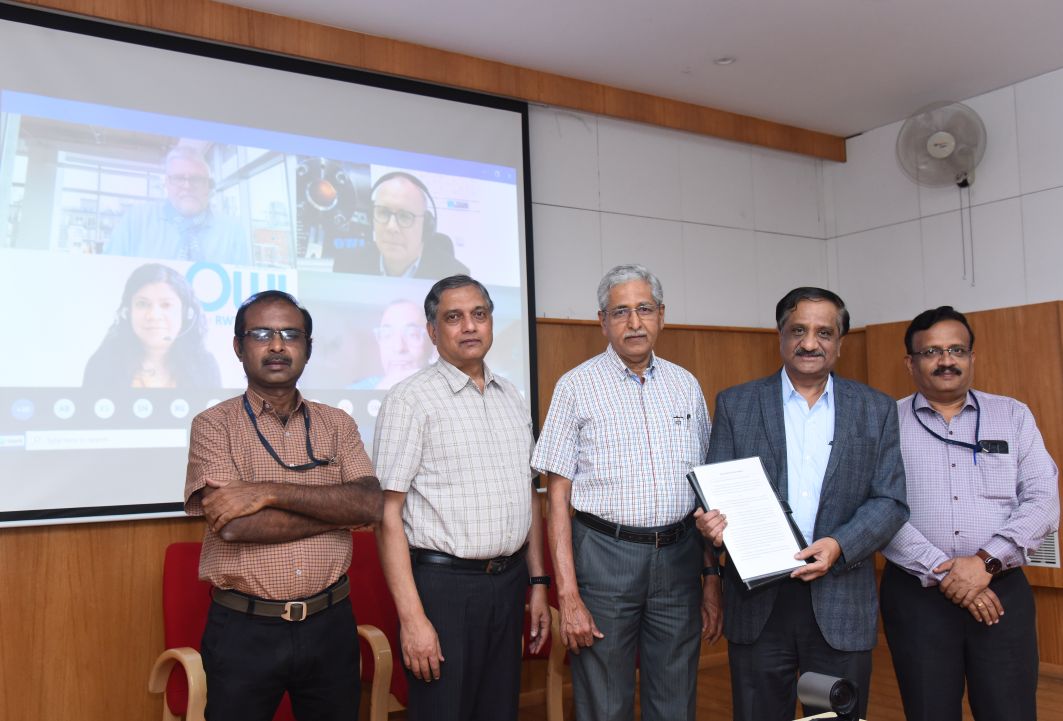 Fünf Männer neben einander stehend blicken in die kamera, im Hintergrund die Projektion weiterer Teilnehmer einer Online-Konferenz.