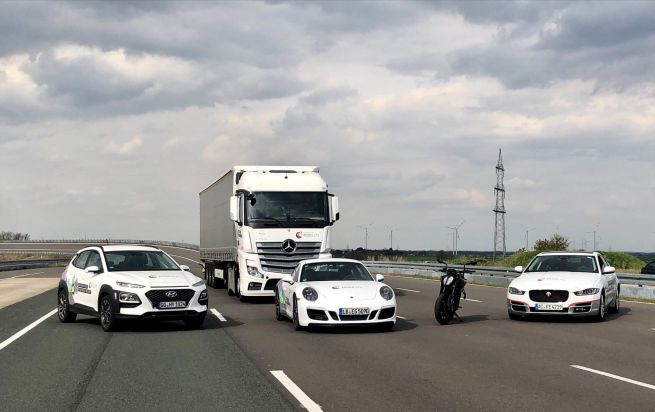 Drei weiße Autos, in Motorrad und ein Lkw stehen auf einer Straße.