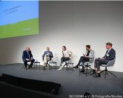 Personen sitzen auf einem Podium im Halbkreis nebeneinander.