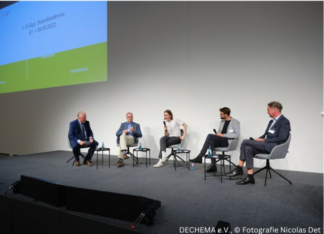 Personen sitzen auf einem Podium im Halbkreis nebeneinander.